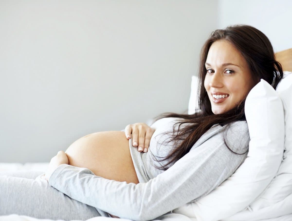 A smiling pregnant woman resting against pillows with her hands on her belly